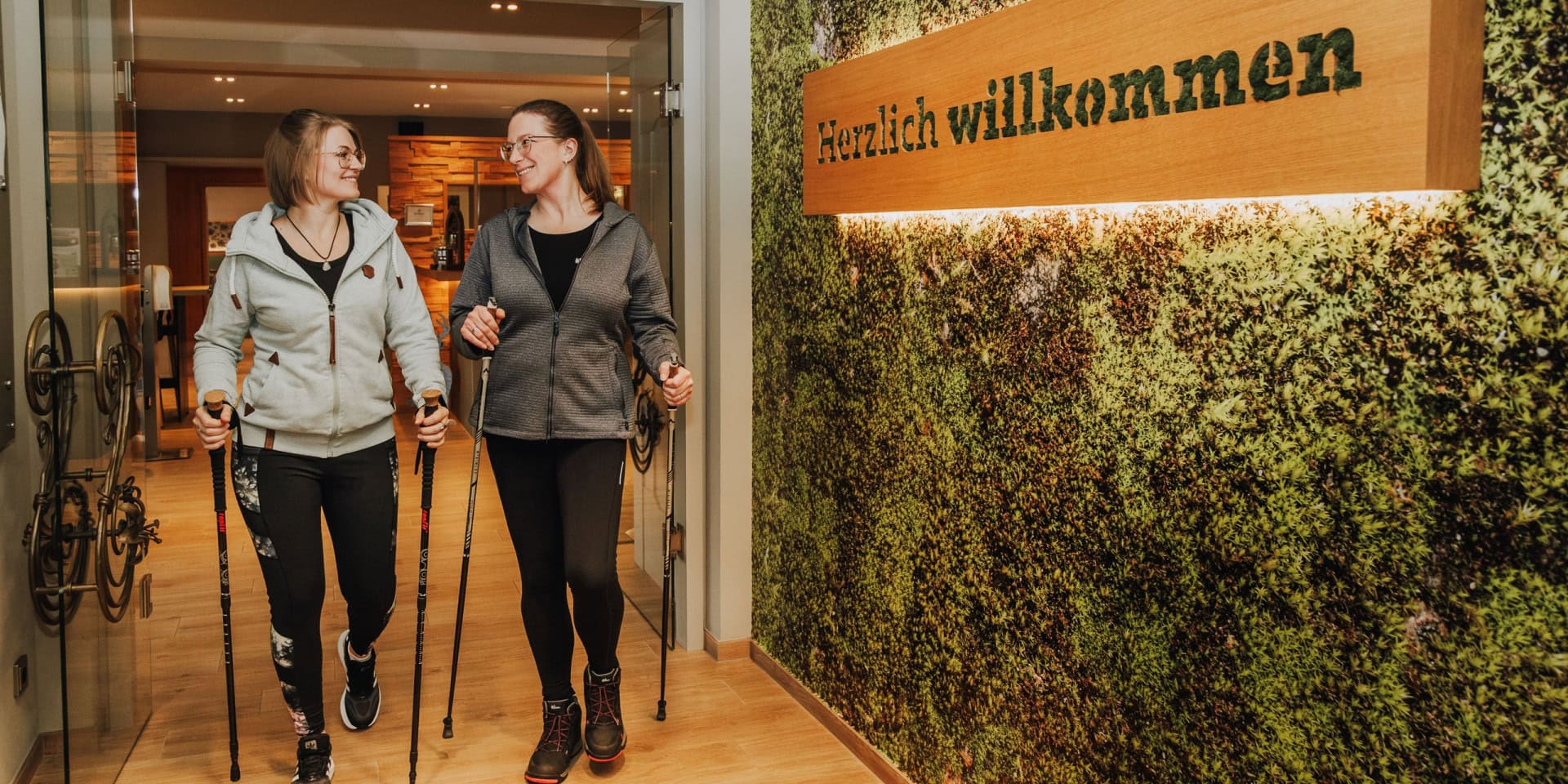 Zwei Frauen mit Nordic-Walking-Stöcken gehen lächelnd durch eine Tür im Hotel, vorbei an einer Mooswand mit Willkommensschild.