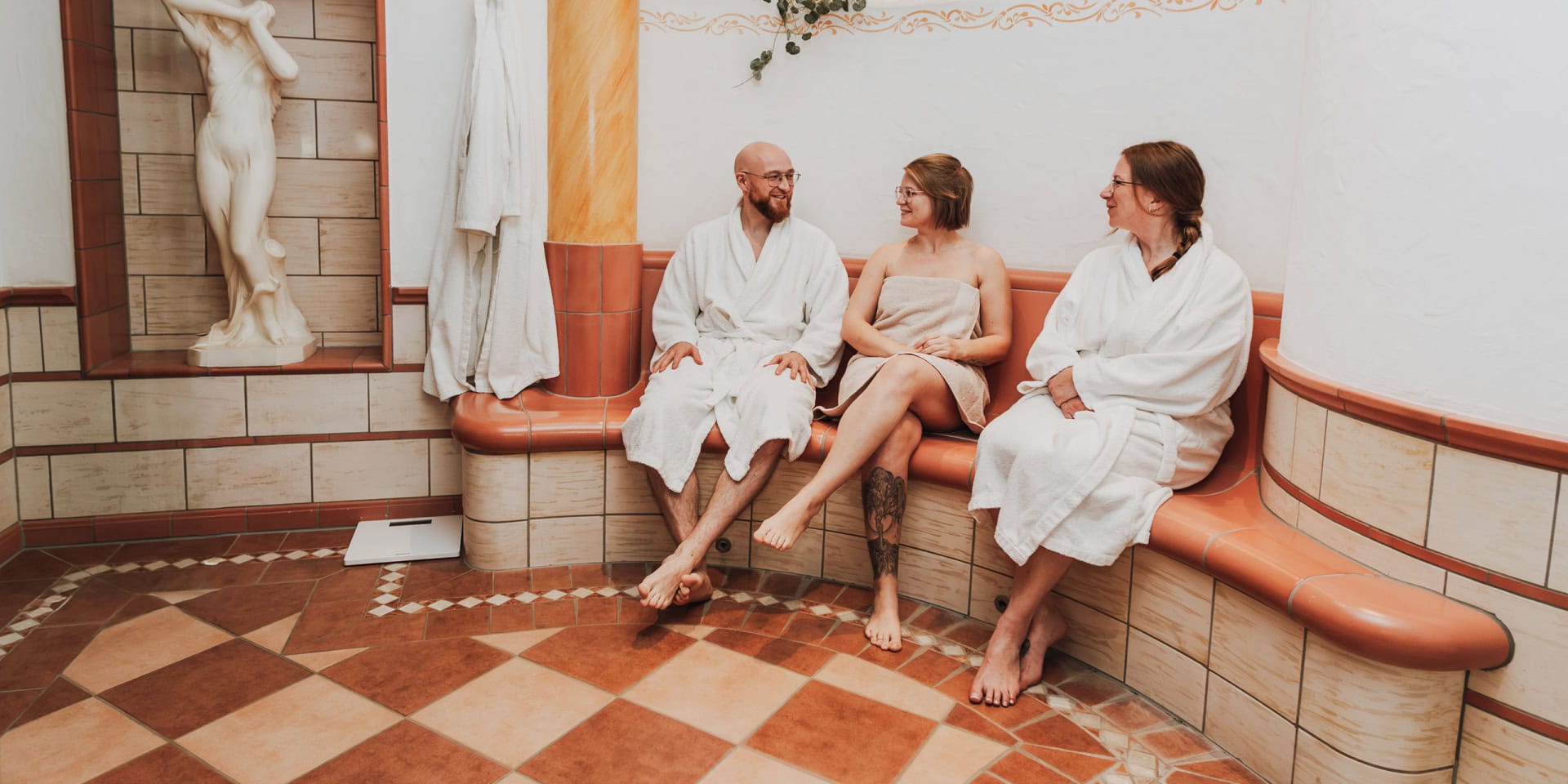 Drei Personen in Bademänteln entspannen sich im Spa-Bereich des Wellnesshotel Panorama in Daun, umgeben von eleganter Dekoration und einer Statue, die Ruhe und Wellness symbolisiert.