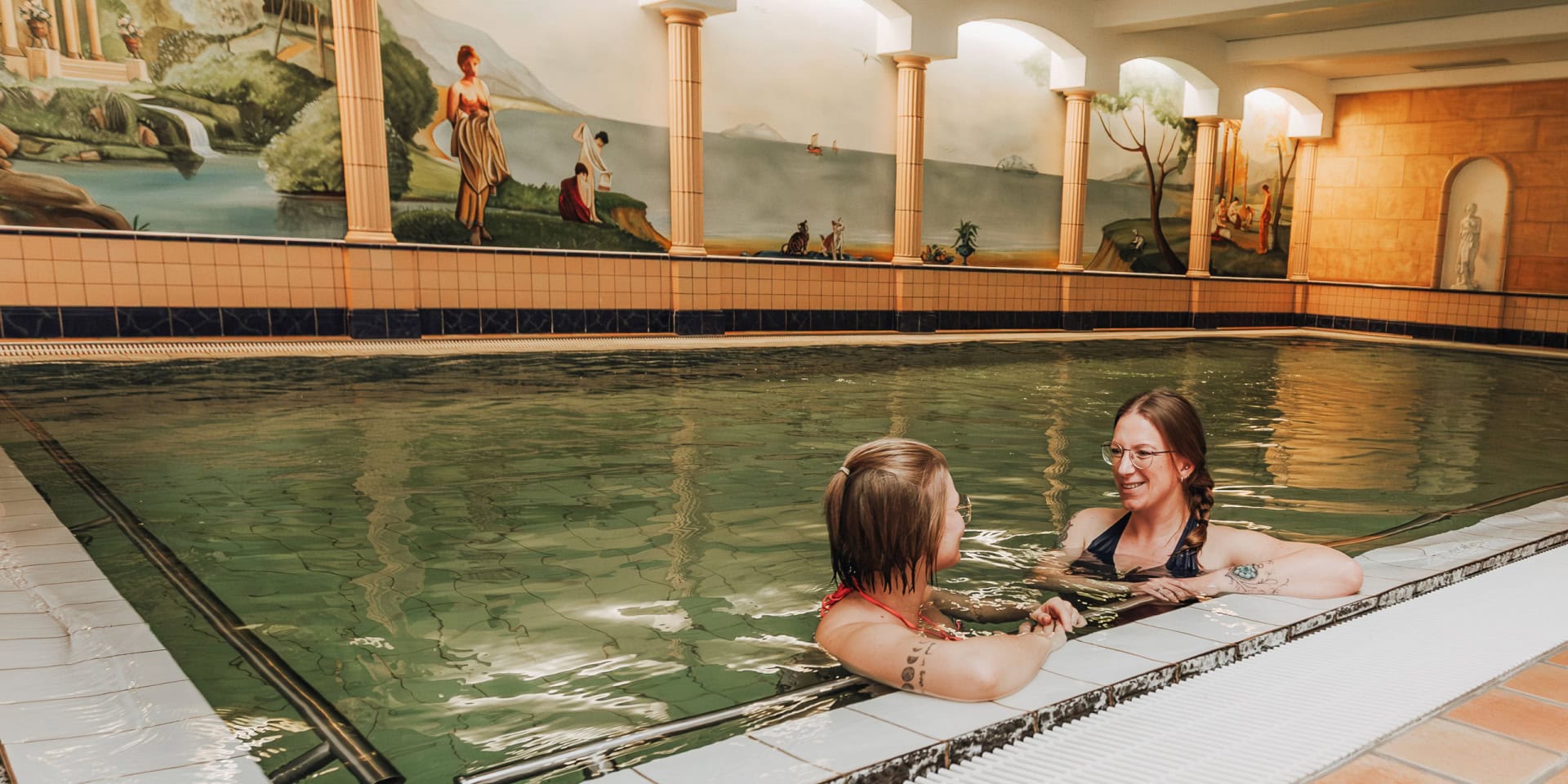 Zwei Frauen entspannen sich im 30 Grad warmen Innenpool des Hotel Panorama in Daun, umgeben von einer kunstvollen Wandmalerei und warmem Licht.